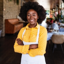 A woman in a yellow shirt and apron stands proudly in front of her restaurant